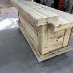 Large unfinished wooden workbench/crate built from vertical planks, on a concrete shop floor with tools in the background.