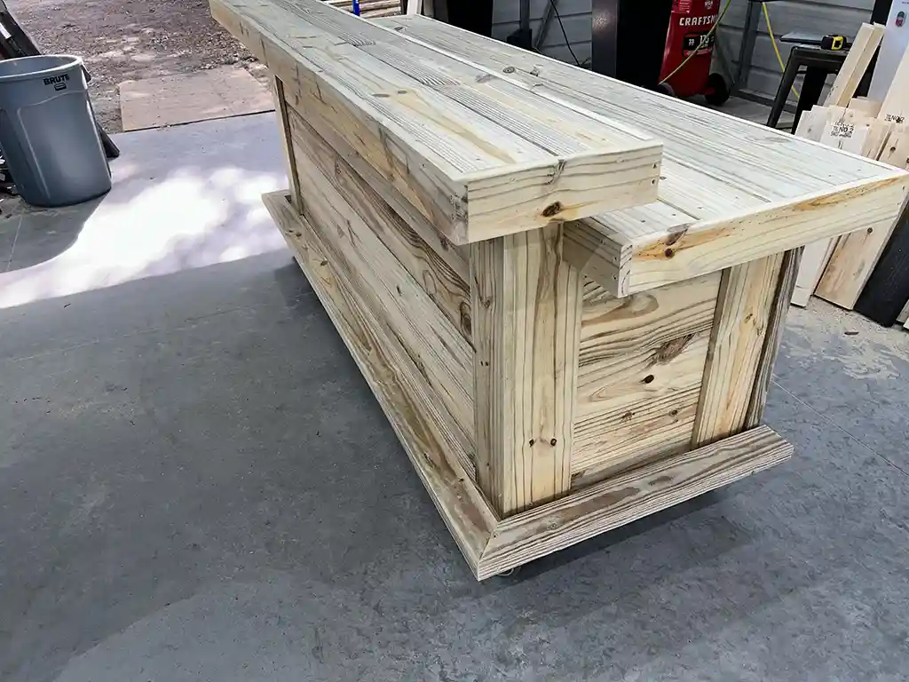 Large unfinished wooden workbench/crate built from vertical planks, on a concrete shop floor with tools in the background.