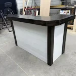 Workbench-style bar counter with a dark wood top and light gray paneled sides in a cluttered workshop.