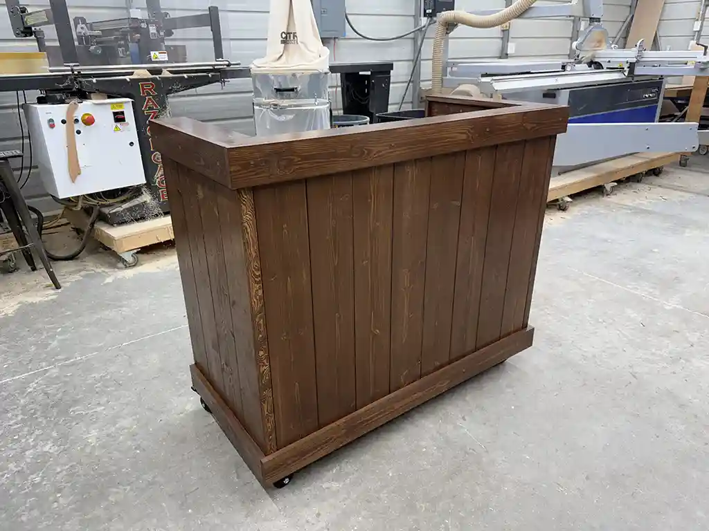 Walnut color Mobile Hostess Stand with 3 shelves 1 Wooden bar or counter on caster wheels in a workshop with machines in the background.