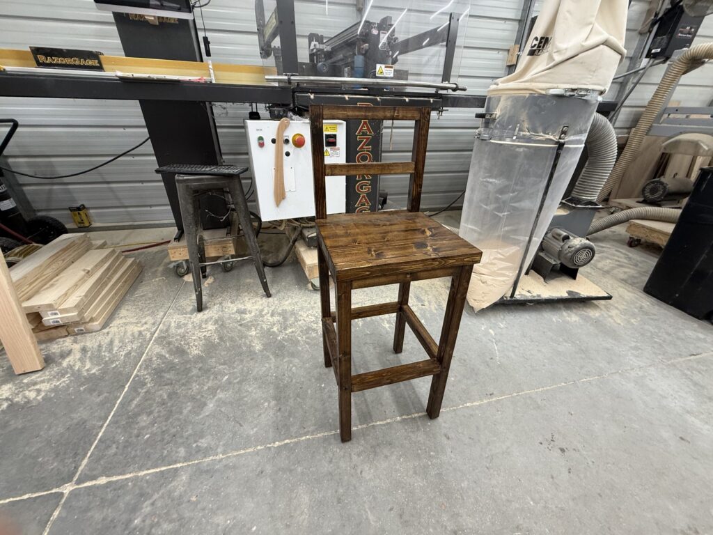 High back barstool provincial color Tall stained wooden stool in a woodworking shop, with a large saw and dust collector in the background and pieces of lumber nearby.